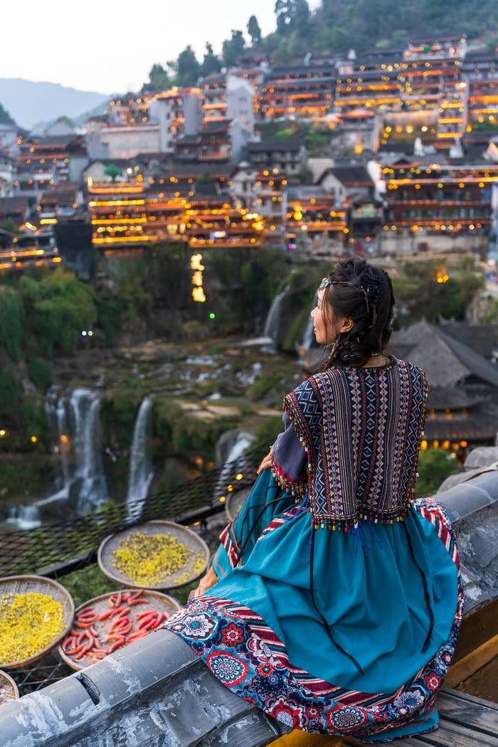 Pueblo tradicional de montaña: Una mujer con vestimenta étnica contempla desde un tejado un pintoresco paisaje de cascadas y casas tradicionales iluminadas al anochecer.