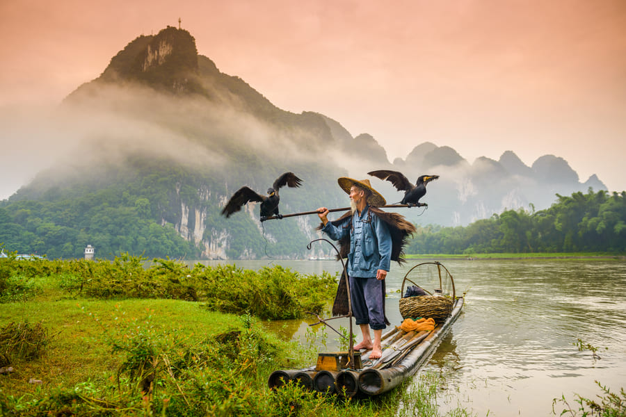 Pescador de cormorán sobre una lancha, en el río Yulong en Yangshuo con montañas de fondo